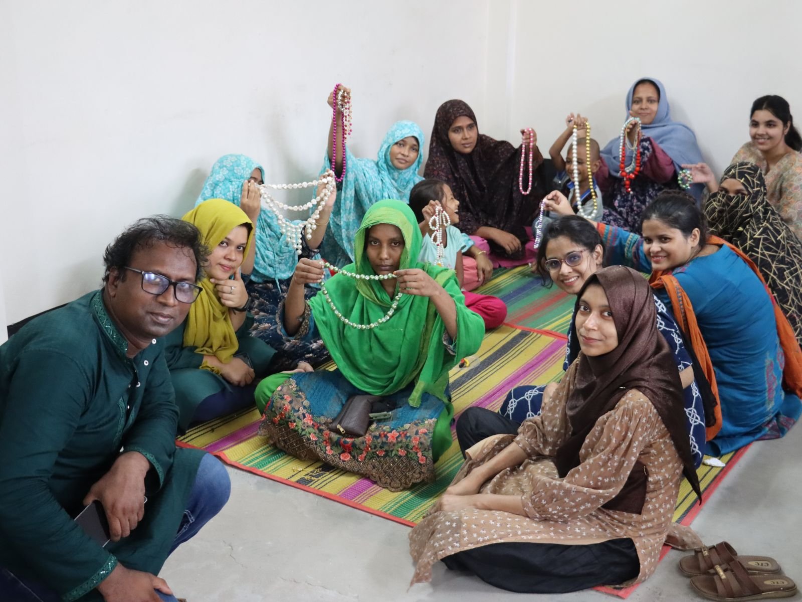 Moner Kaj makers sitting together on a mat, holding up the beaded necklaces they have just strung