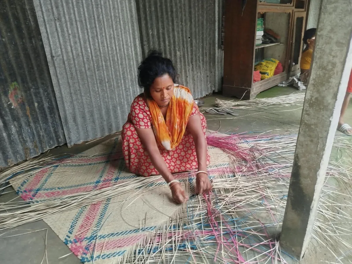A woman weaving a shital pati mat on the floor, threading pink and blue strands of cane through the pattern