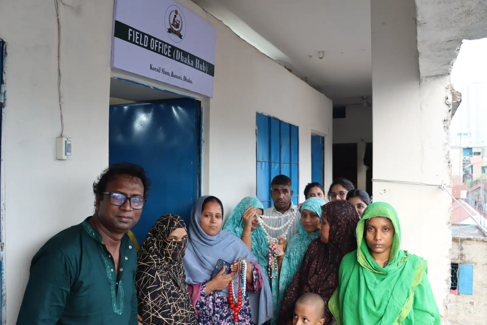 Artisans and staff outside the Moner Kaj field office in Korail, Dhaka, holding the bead necklaces they made