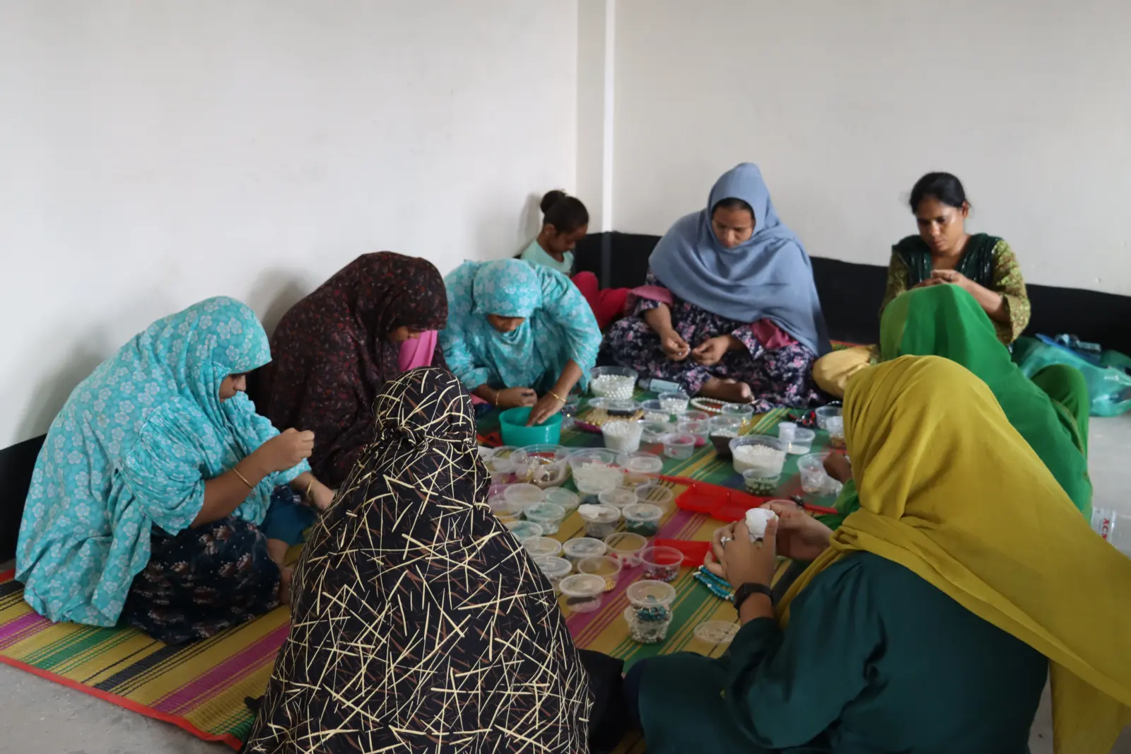 Women sitting on a woven mat, sorting beads from small tubs and threading jewellery