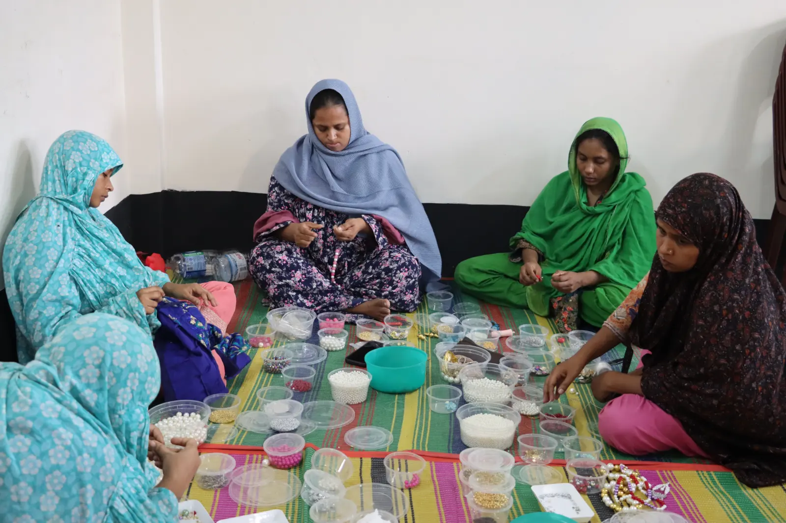 Women sitting together on a woven mat, threading beads from small tubs in a group session